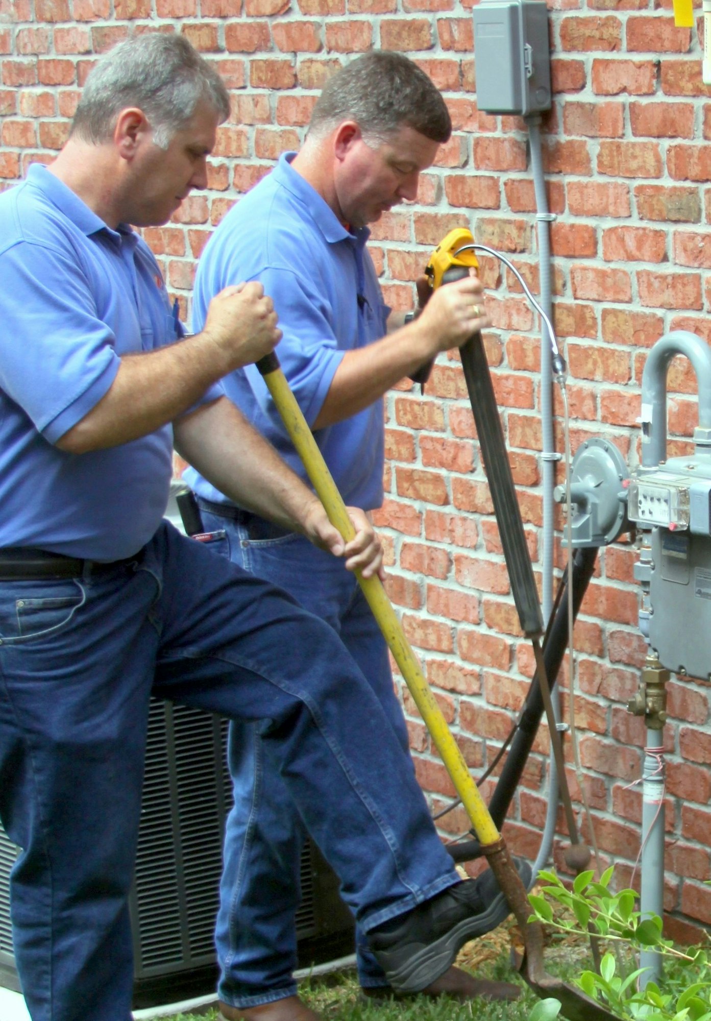 Two men in blue shirts work on a gas meter outside a brick building.