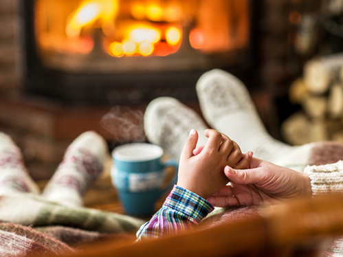 Cozy scene by a fireplace, showing feet in socks, a cup of coffee, and someone holding a child's hand.