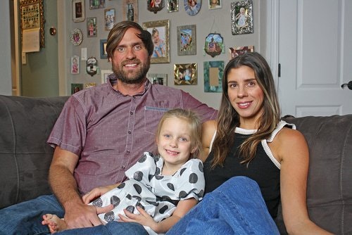 A family of three sitting on a couch; a man, woman, and a young girl, with framed photos on the wall behind them.