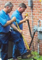 Two people in blue shirts working on a gas line or meter by a brick wall, using hand tools.