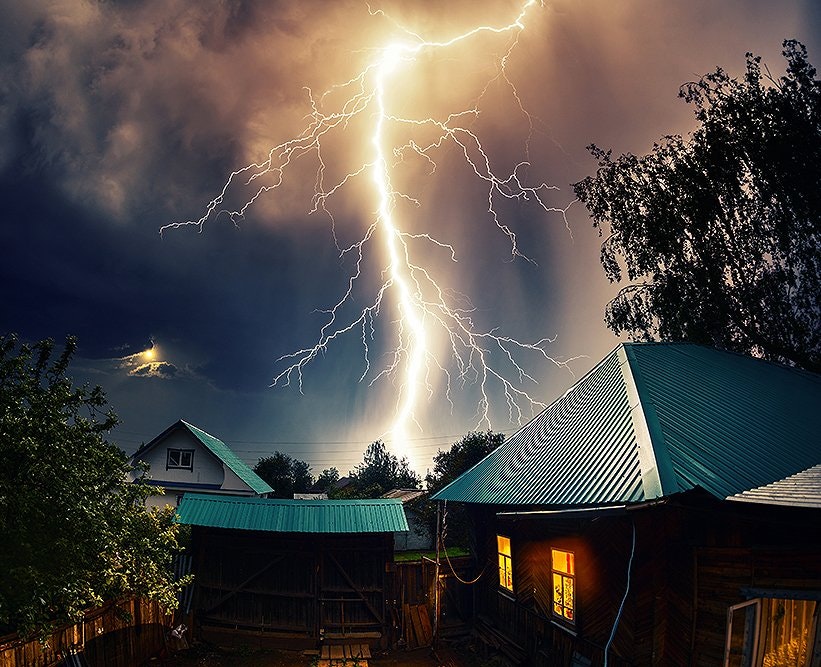 Powerful lightning bolt striking behind houses at night with lit windows, dramatic sky.