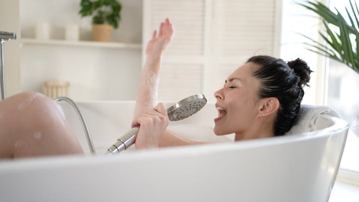 Person singing in the bathtub, using a showerhead as a microphone, with a plant in the background.