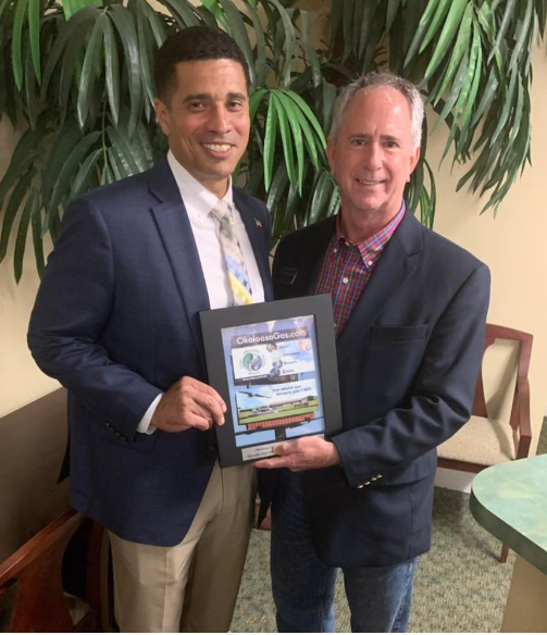 Two men in suits smiling, holding a framed certificate from Okaloosa Gas in a room with plants.