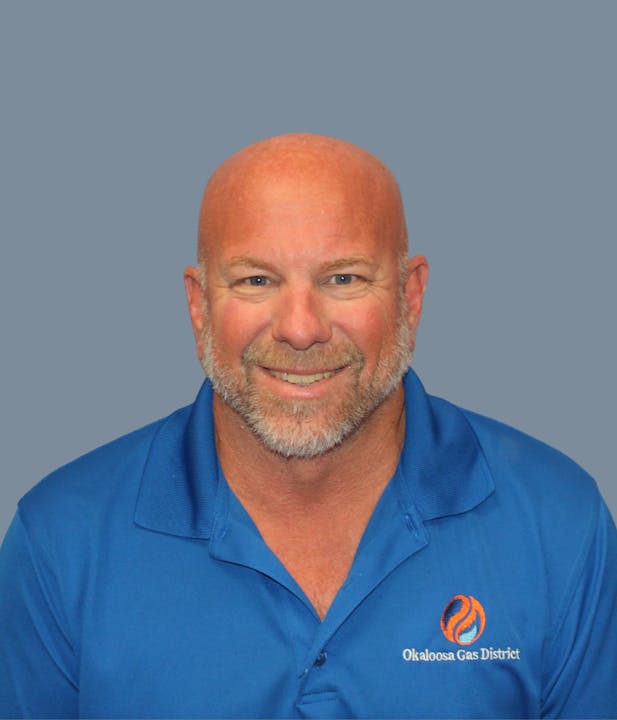 The image features a smiling man in a blue polo shirt with the logo of Okaloosa Gas District, against a gray background.