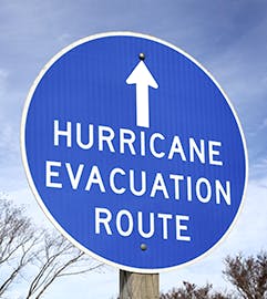 A blue sign indicating "Hurricane Evacuation Route" with an arrow pointing up, set against a rural landscape.