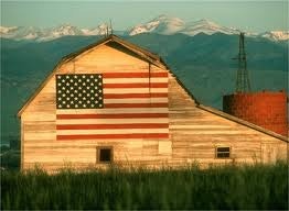 A rustic barn features a painted American flag, set against a backdrop of snow-capped mountains. Green fields surround the structure.