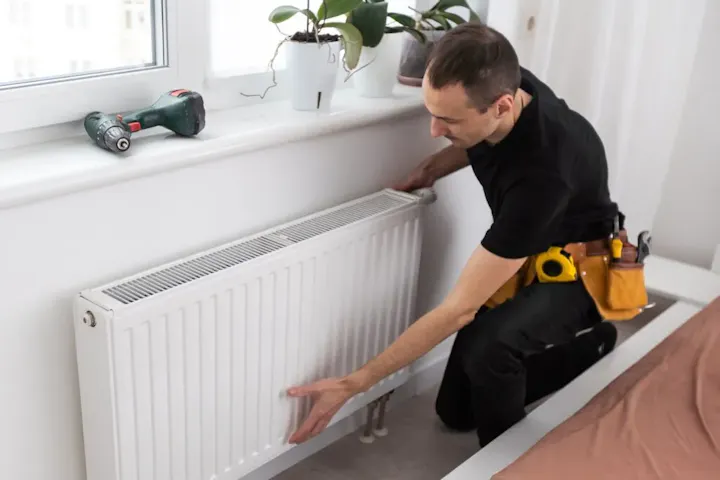 Man in black shirt installing or adjusting a radiator next to a window. A drill and plants are on the windowsill.