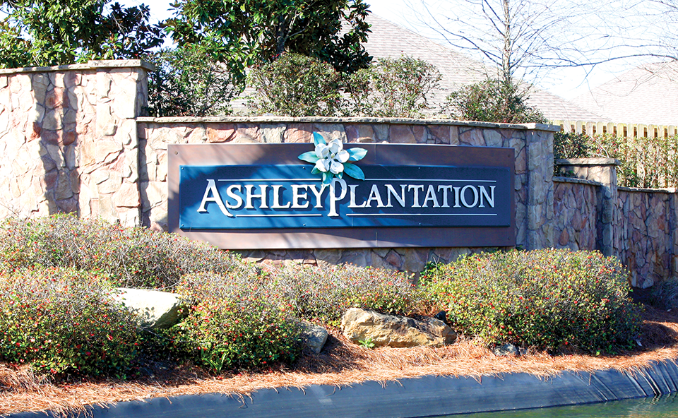 A sign reading "Ashley Plantation" on a stone wall, surrounded by shrubbery.