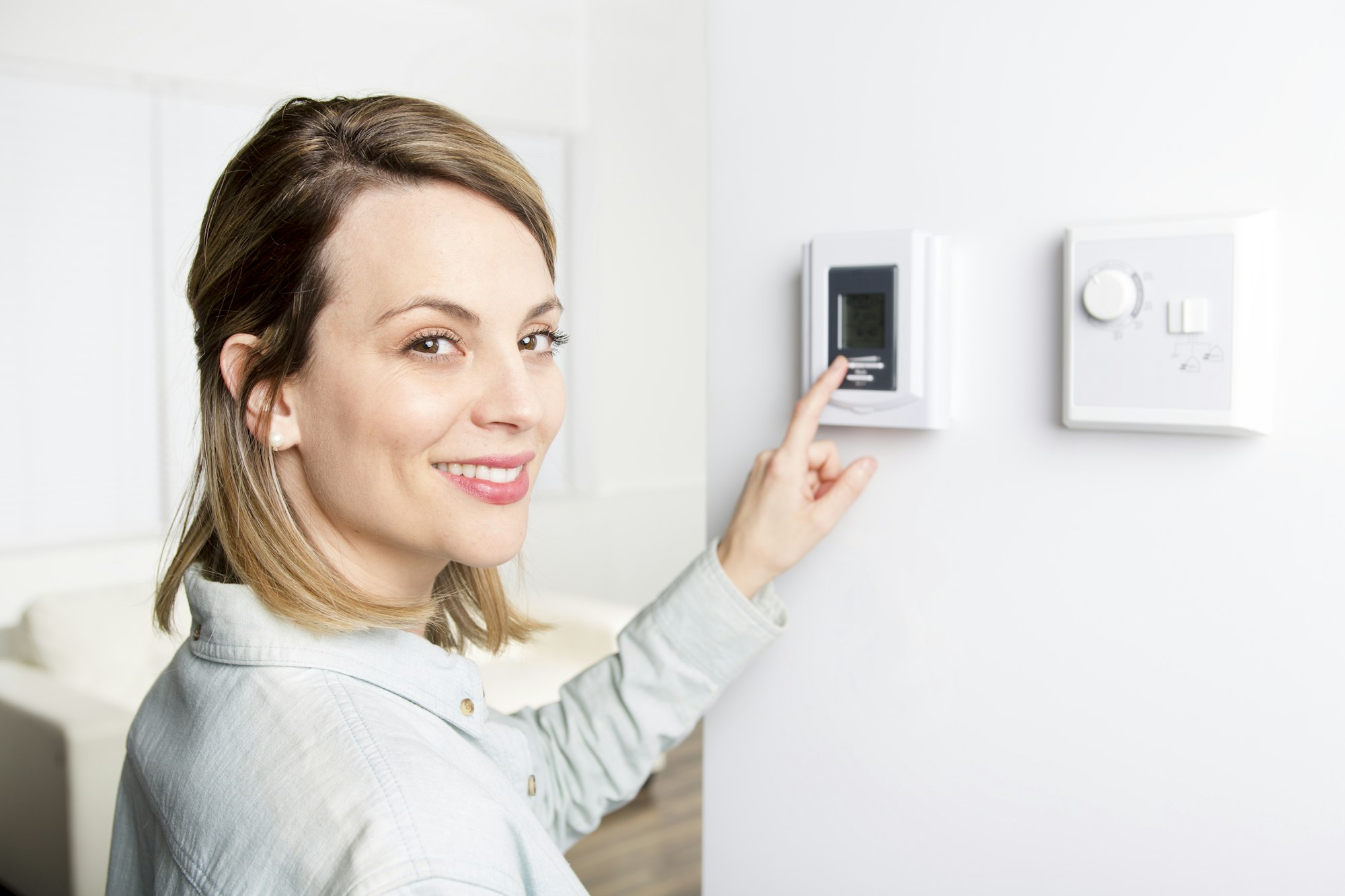 A person interacts with a thermostat on the wall, smiling, in a bright indoor setting.
