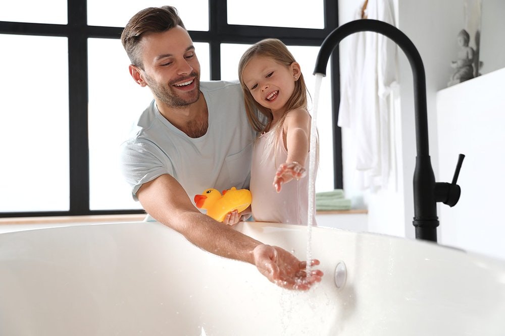 Man and girl playing with water in bathtub; girl holds a rubber ducky; both smiling.