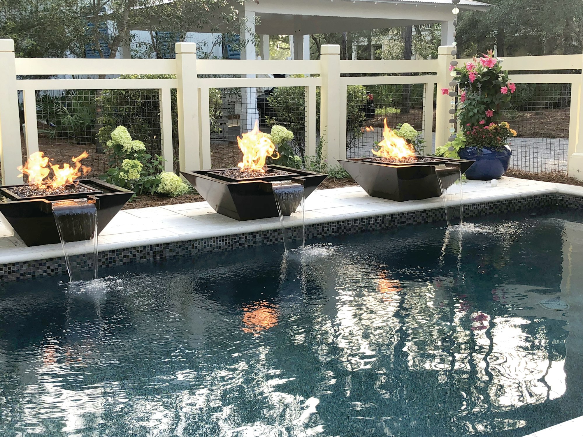 A serene pool setting featuring three stylish fire bowls with flames and water features, surrounded by greenery and flowers.