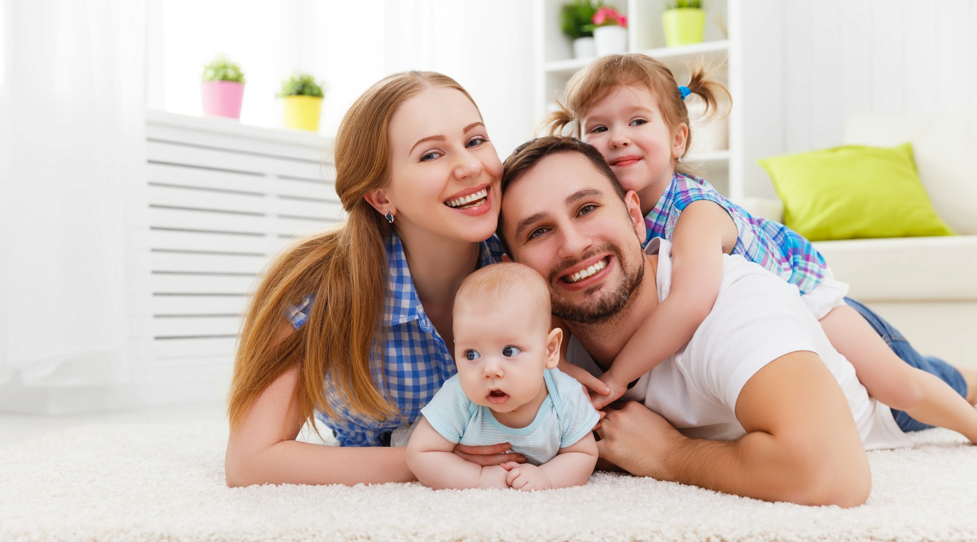 A smiling family of four lying on the floor: two parents, a baby, and a young child.