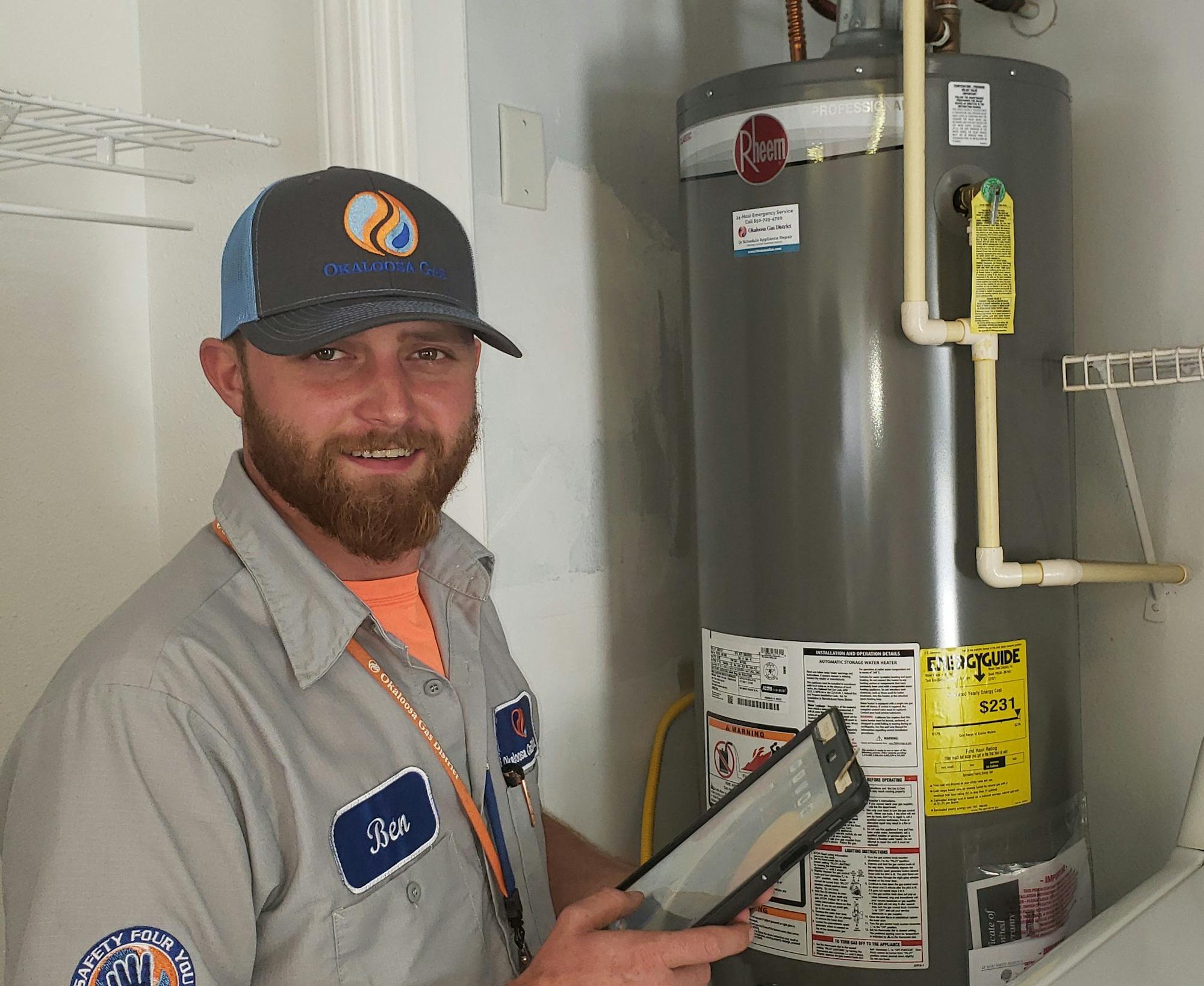 A man holding a tablet stands next to a water heater in a utility room with a dryer.