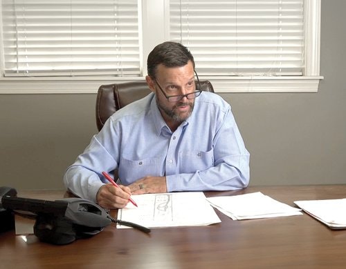 A man seated at a desk, holding a red pen, with documents in front and a telephone nearby.