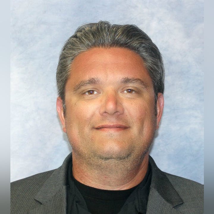 A person with short gray hair and a slight smile is facing forward against a light background.