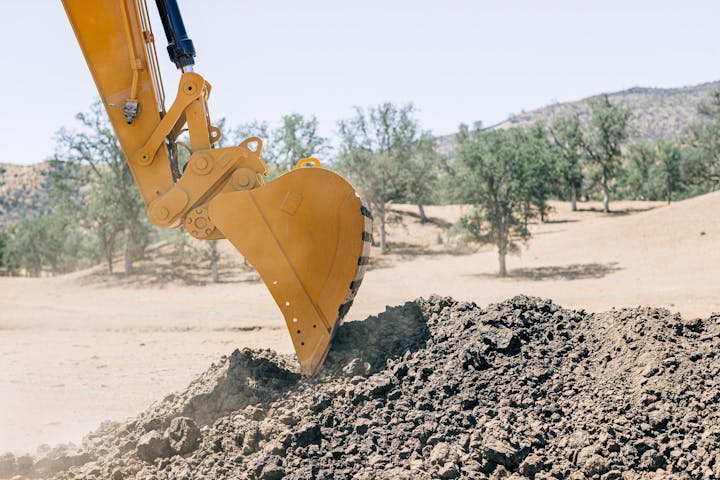An excavator's bucket digging into a pile of dirt in a desert landscape with trees in the background.