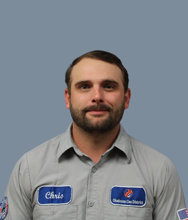 The image shows a man with a beard wearing a uniform with patches that say "Chris" and "Okaloosa Gas District." Background is light blue.