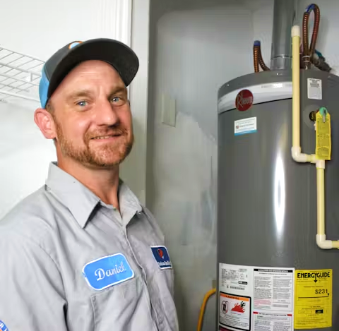 A smiling maintenance technician stands next to a water heater, wearing a uniform and cap, ready for service.