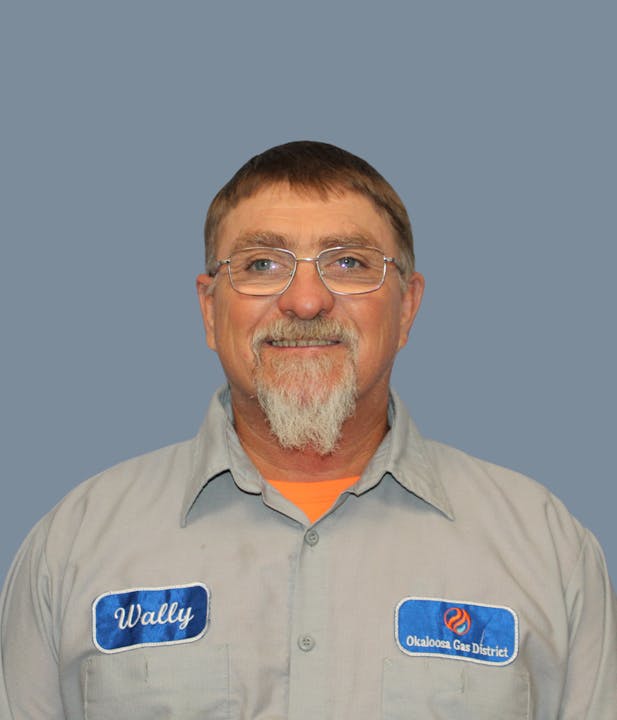 The image shows a smiling man with glasses, wearing a work shirt with "Wally" and "Okaloosa Gas District" badges.