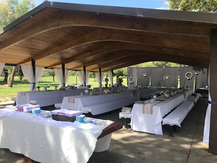 The image shows a decorated outdoor pavilion with long tables, white coverings, and floral arrangements, likely set for an event.