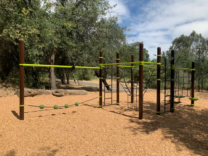 The image shows a modern playground with climbing structures, ropes, and green balls, set in a natural outdoor area.