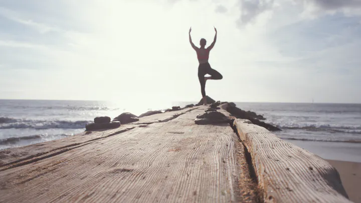 A person practices yoga on a wooden pier by the ocean, silhouetted against a bright, cloudy sky. Serenity and balance captured.