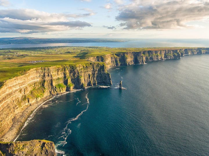 Cliffs meeting the sea under a cloudy sky; lush green landscape on top.