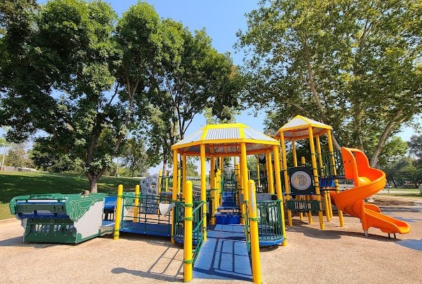 A colorful playground structure with slides, climbing areas, and surrounded by trees in a sunny park setting.