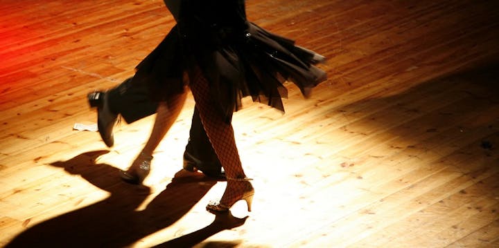 A close-up of two dancers' legs in motion on a wooden floor, showcasing elegant footwear and flowing garments under soft lighting.