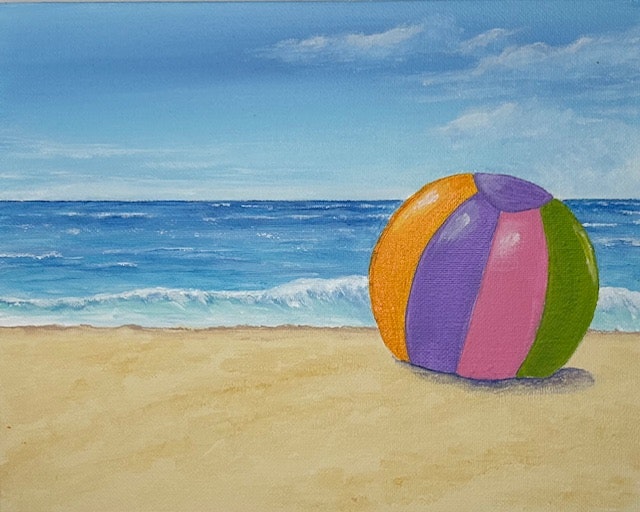 Colorful beach ball on sandy shore with waves in the background under a blue sky.