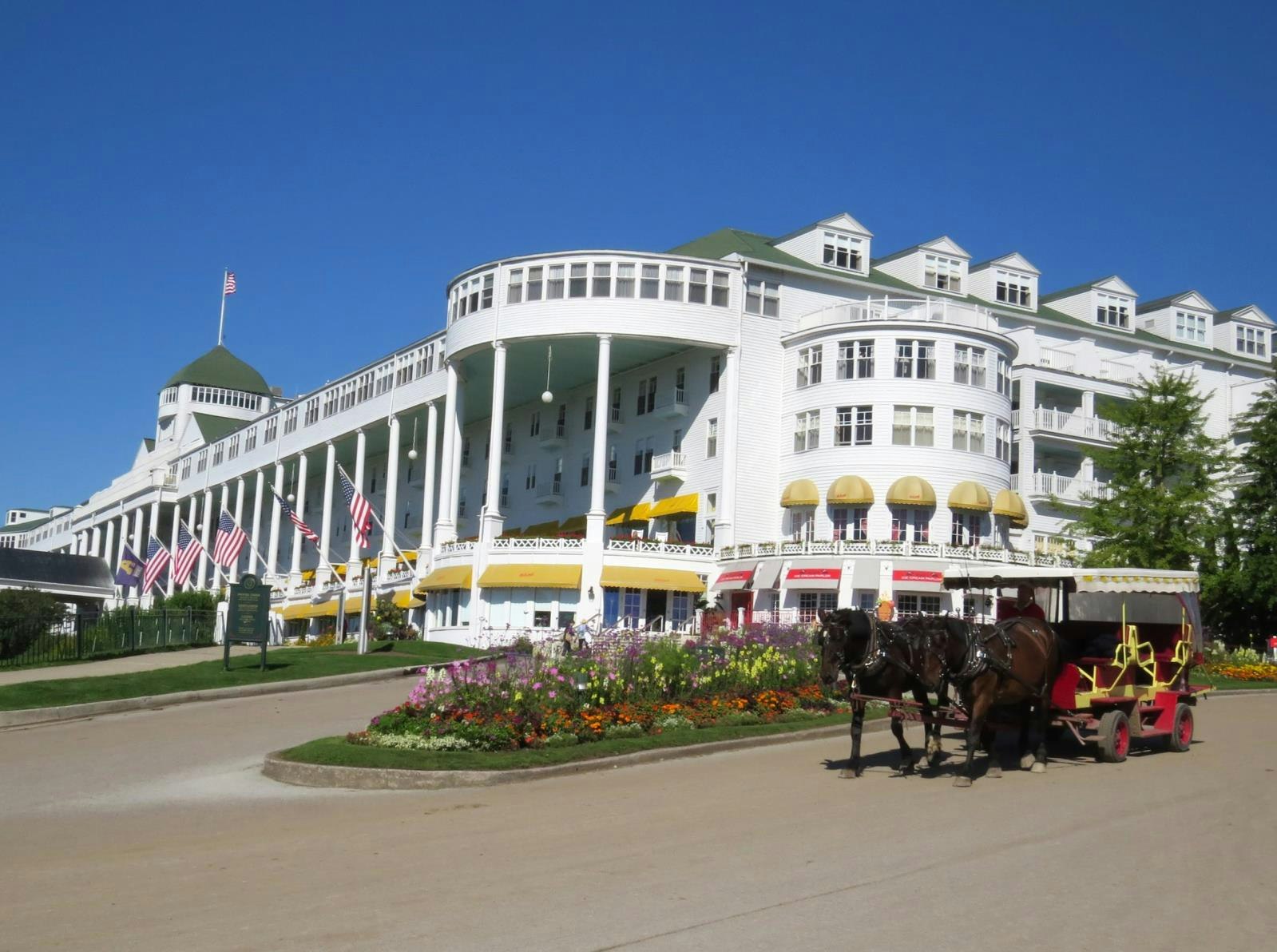 A large white hotel with American flags, surrounded by flowers and a horse-drawn carriage in front.