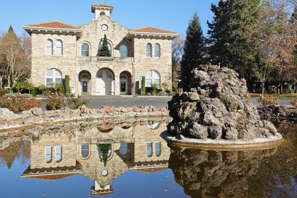 A stone building with a clock tower reflects in a pond, featuring a rocky fountain and surrounded by trees and greenery.