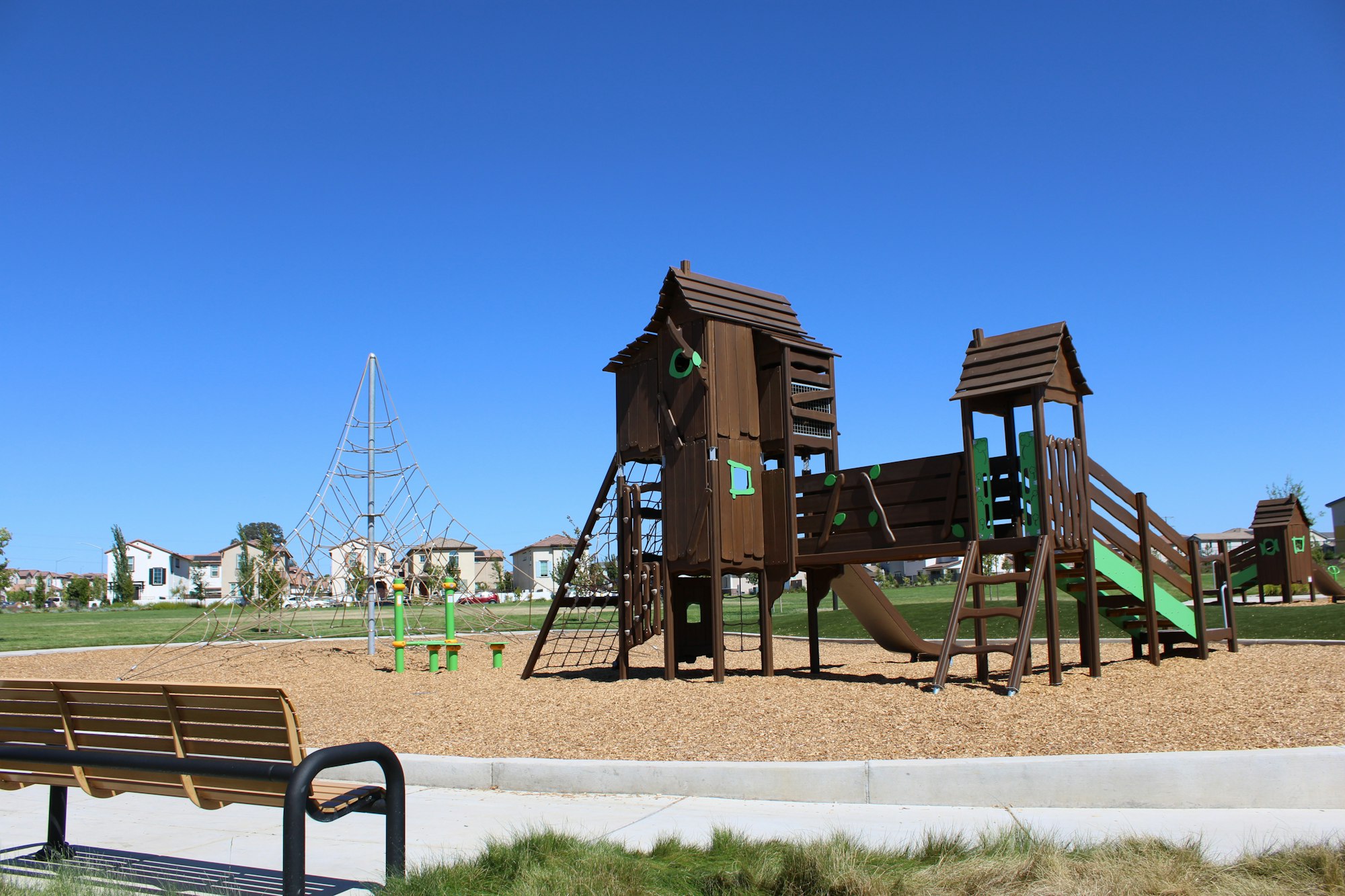 A playground featuring a wooden play structure, climbing web, and benches, set against a clear blue sky and green grass.