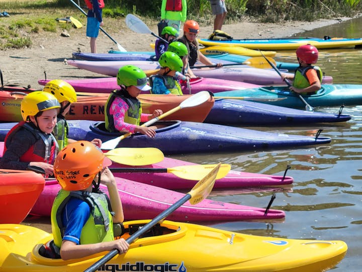Colorful kayaks and children wearing helmets and life jackets preparing for kayaking.