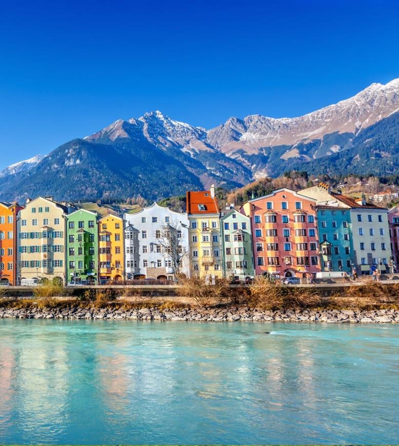 Colorful houses by a river with a backdrop of snow-capped mountains under a clear blue sky.