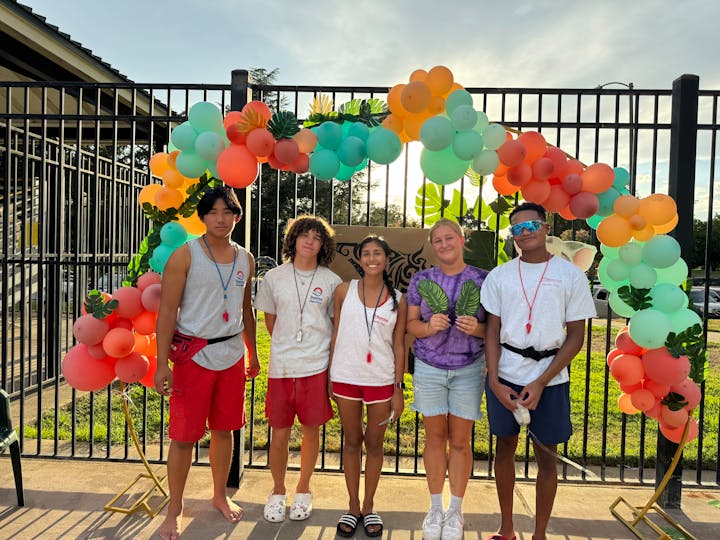 A group of lifeguards poses in front of a colorful balloon arch at a pool, smiling and holding props, showcasing a fun atmosphere.