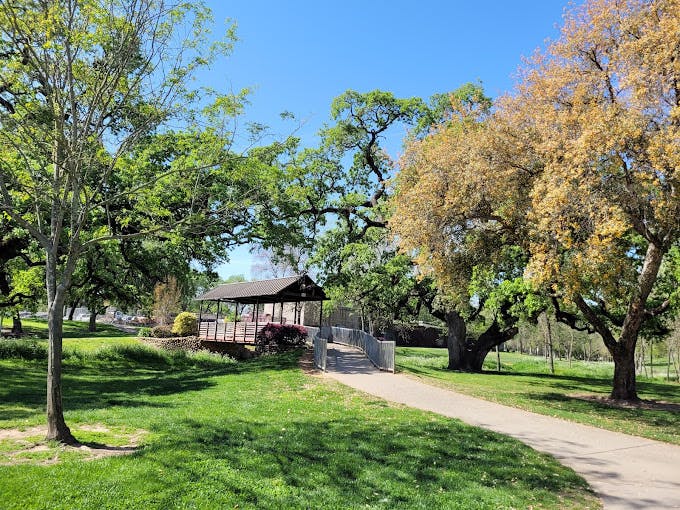 A park with green grass, trees, a gazebo, and a walking path, under a clear blue sky.