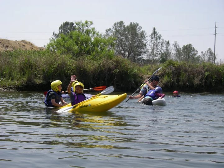 A group of people enjoying kayaking and swimming in a river on a sunny day, wearing life jackets and helmets.