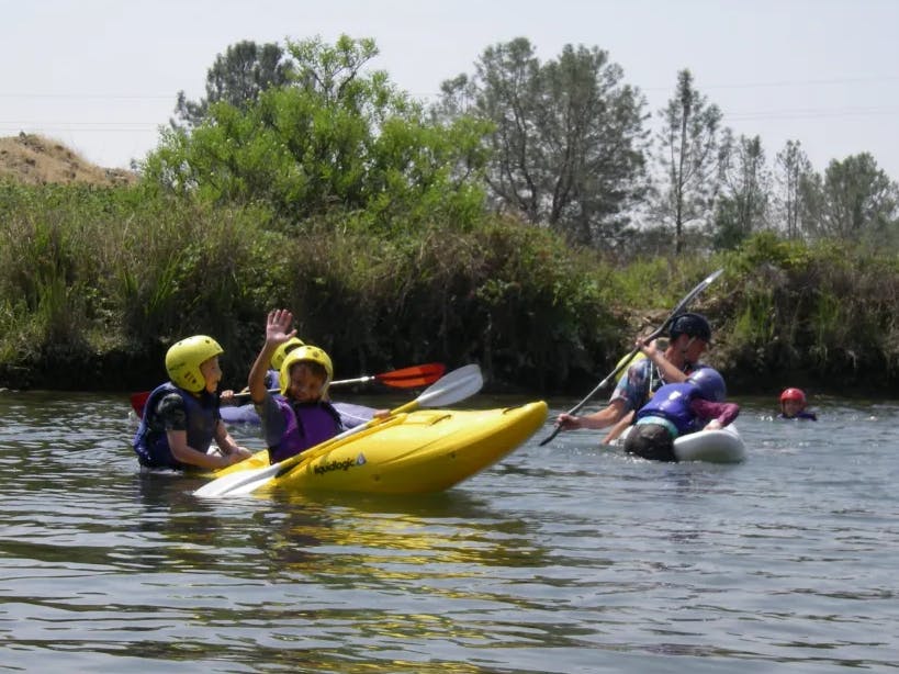 A group of people enjoying kayaking and swimming in a river on a sunny day, wearing life jackets and helmets.
