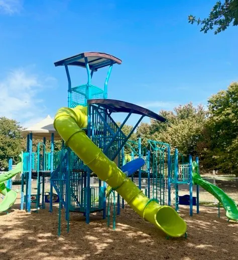 A colorful playground featuring a tall structure with a spiral slide and green slides, set against a blue sky and trees.