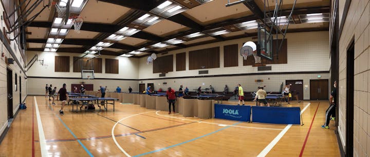 An indoor sports facility with multiple table tennis tables and people playing, alongside basketball hoops and seating areas.