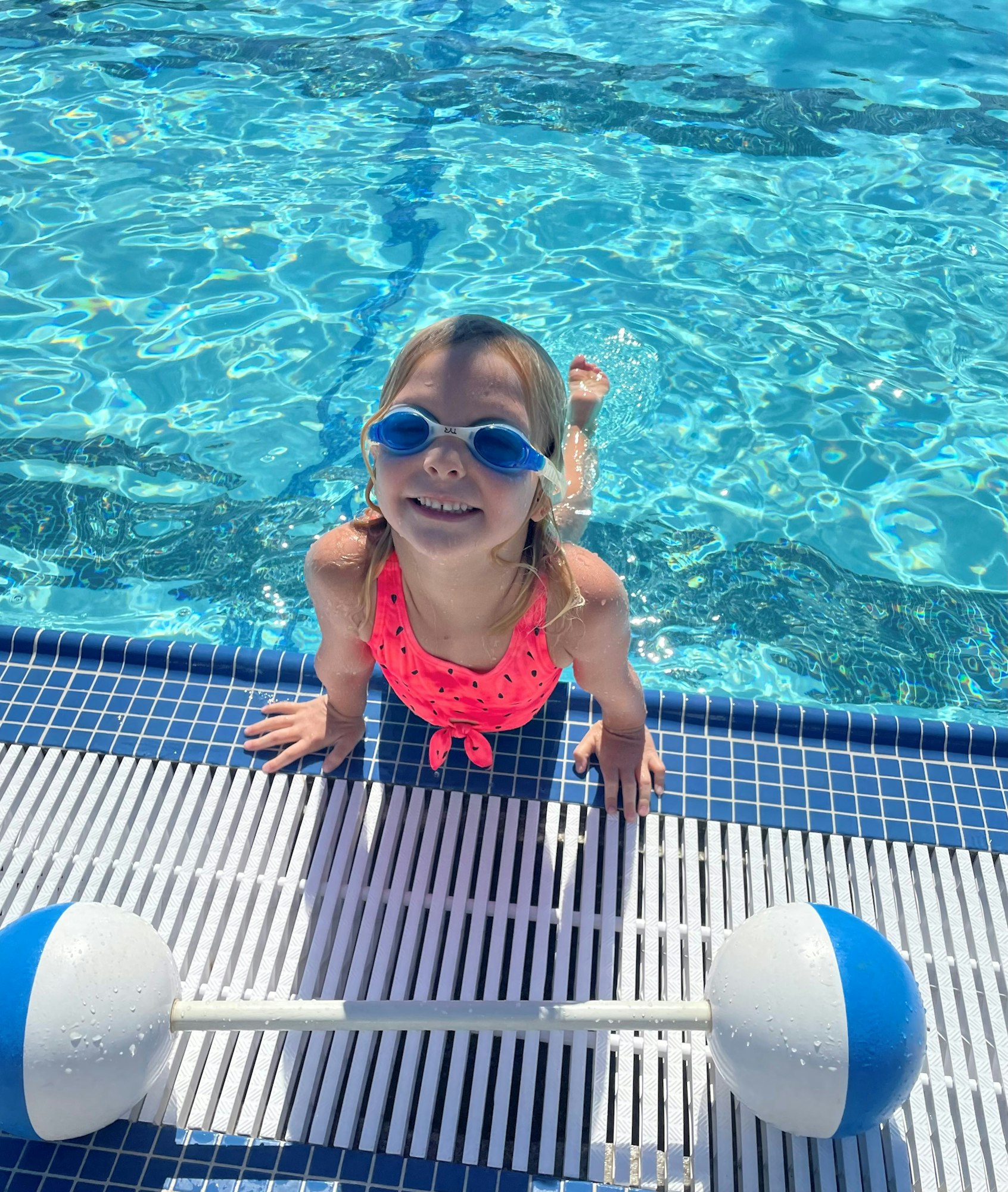 A smiling child wearing goggles in a pool, with a floating pool buoy.