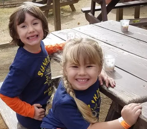 Two smiling kids in matching blue t-shirts pose together at a picnic table, with cups in front of them.
