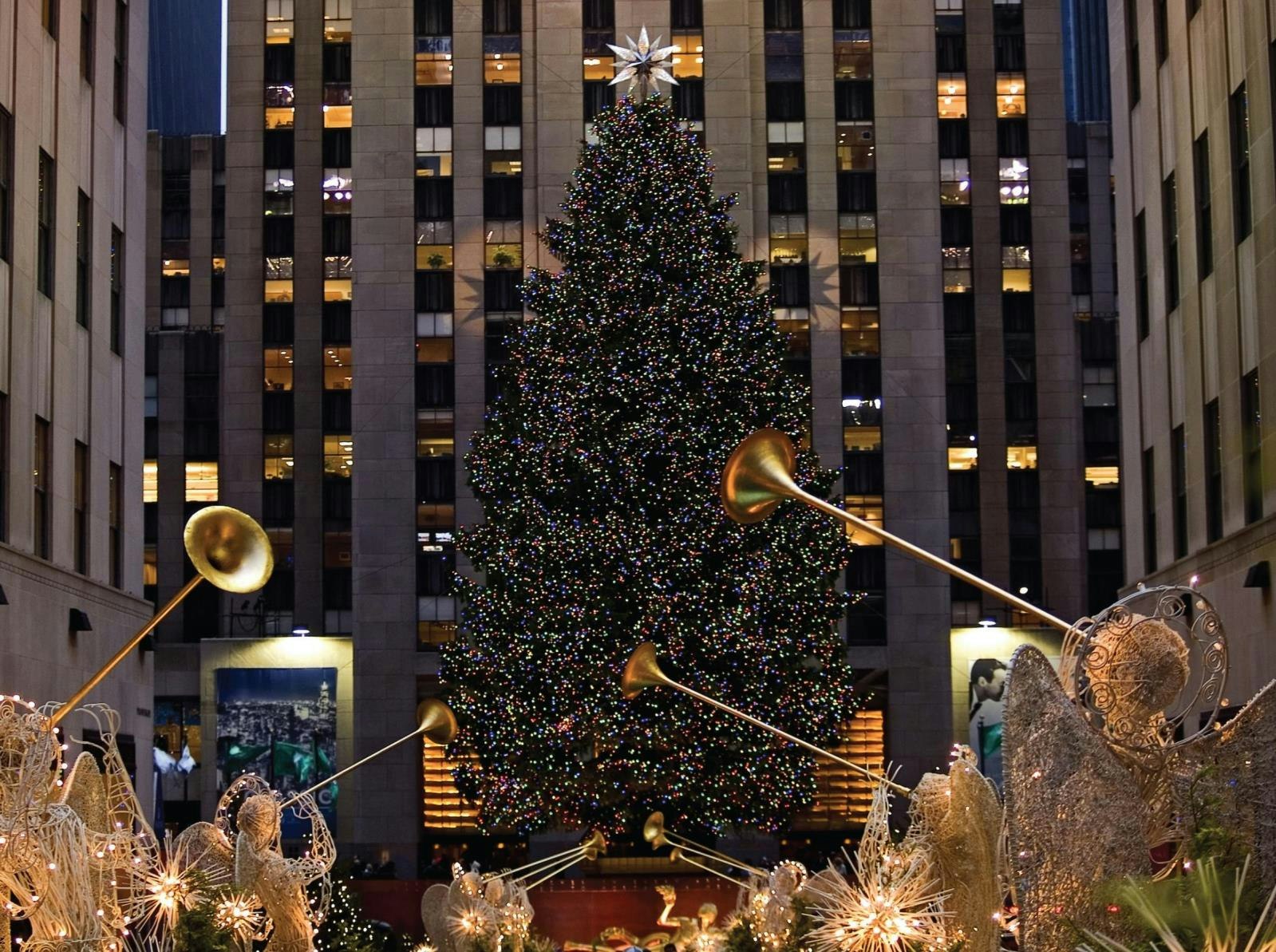 A large, brightly-lit Christmas tree with trumpet-playing angel decorations in an outdoor setting.