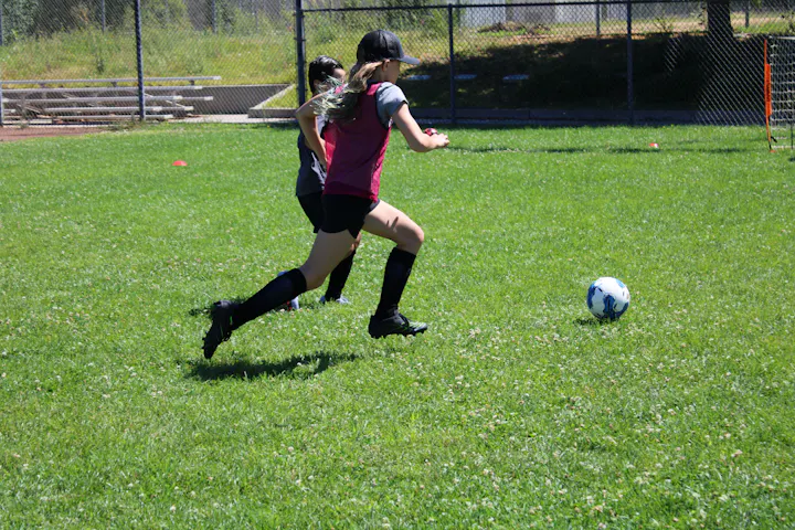 Two girls are playing soccer on a green field, with one actively running towards the ball.
