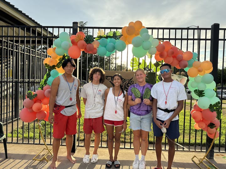 Five individuals pose together in swimwear near a colorful balloon arch, likely at a summertime event or pool setting.