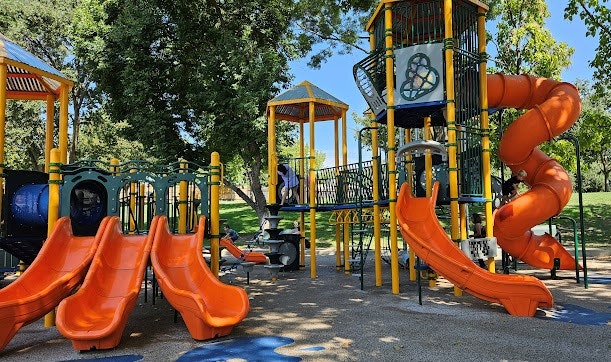 A colorful playground with multiple slides and climbing structures, surrounded by trees and an open grassy area.