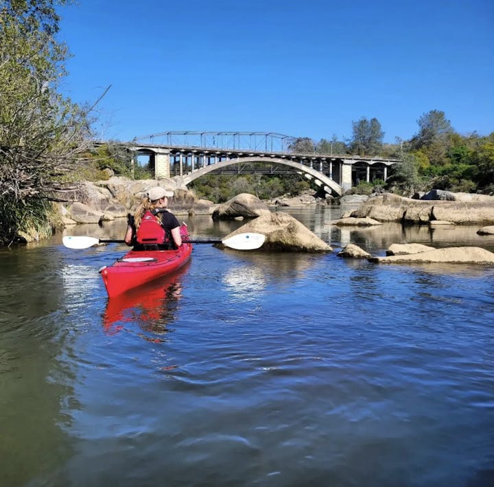 A person in a red kayak paddles on a calm river, with a bridge in the background and rocky shores under a clear blue sky.