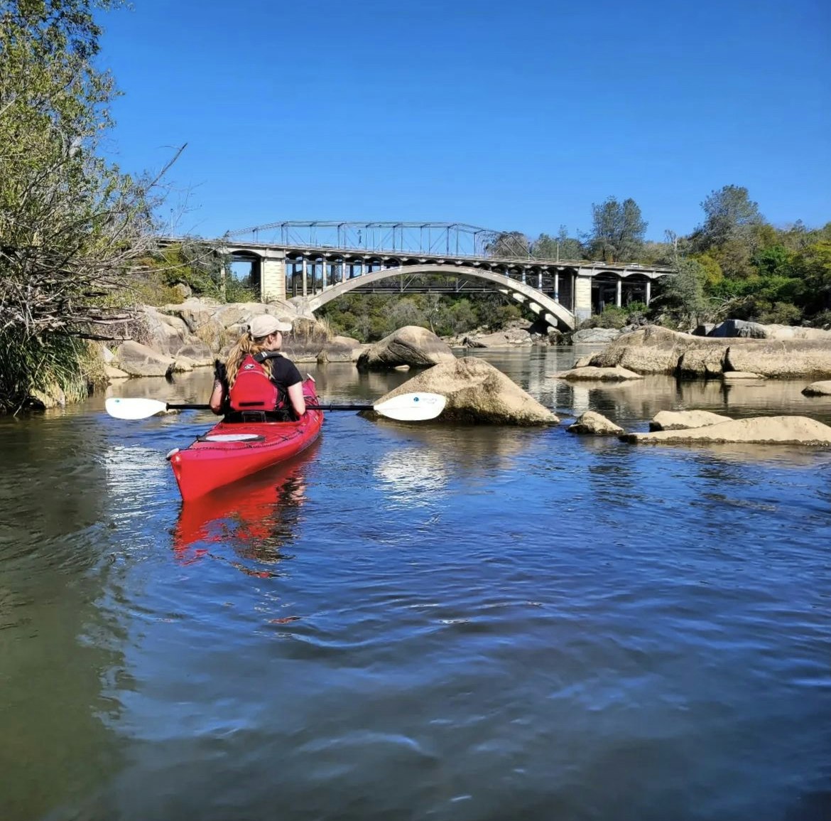 A person in a red kayak paddles on a calm river, with a bridge in the background and rocky shores under a clear blue sky.