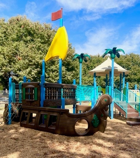 A colorful playground featuring a pirate shipstructure, complete with a yellow sail, surrounded by trees and a play area.